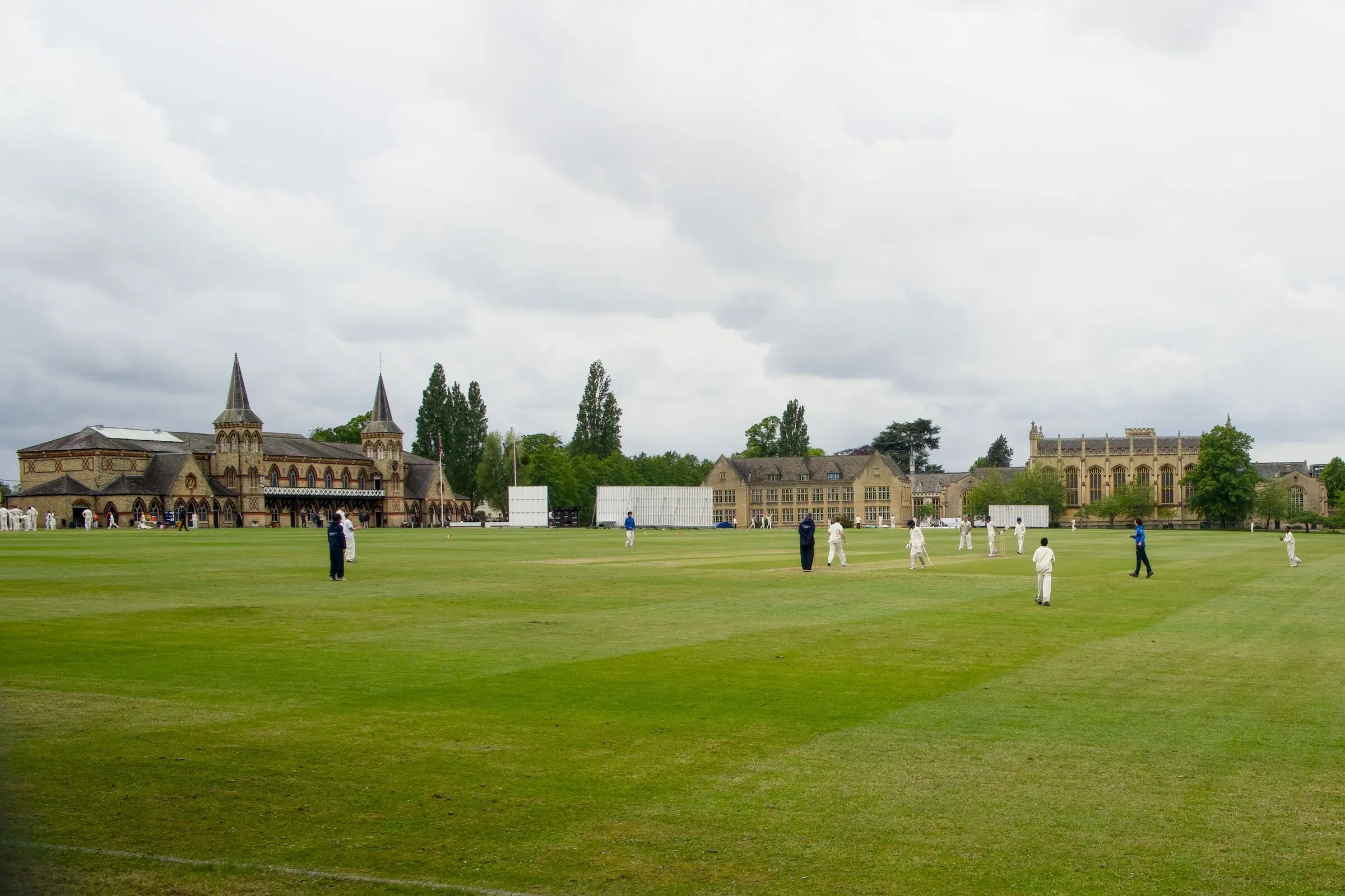 Cricket in progress on a large mown outfield with collegiate buildings behind under overcast cloud